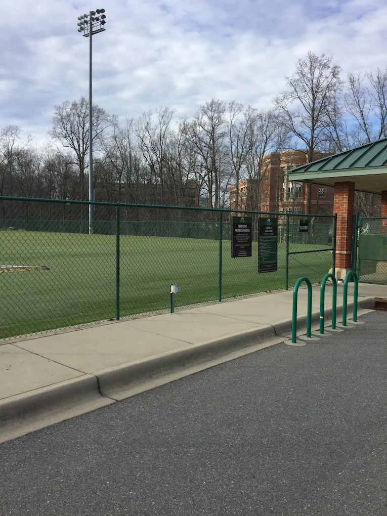 Sports field entrance with green chain-link fence, tall light pole, and bike racks